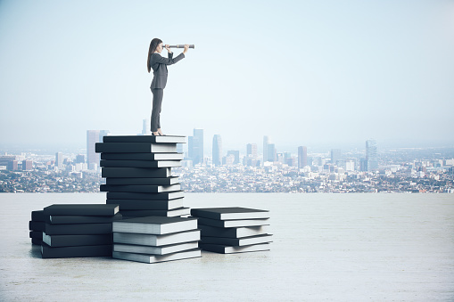 Young businesswoman with telescope standing on book pile. City skyline background. Future and vision concept