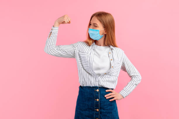 Happy healthy strong young woman in a medical protective mask on her face, showing biceps on her arm, on an isolated pink background Happy healthy strong young woman in a medical protective mask on her face, showing biceps on her arm, on an pink background immune system photos stock pictures, royalty-free photos & images
