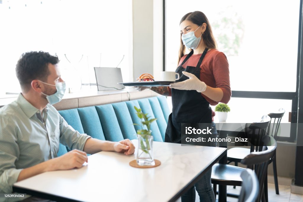 Waitress Serving Order To Customer In Cafe Man looking at waitress serving during coronavirus outbreak in restaurant Restaurant Stock Photo