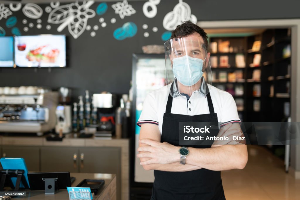 Man With Face Shield And Mask Working In Cafe Portrait of worker wearing face shield and mask standing in cafe with arms crossed Coronavirus Stock Photo