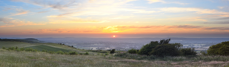 Panoramic view from Ditchling Beacon during sunset, with a dramatic cloudy sky and grass meadows in the foreground. Located on the South Downs, Ditchling Beacon is the highest point in East Sussex.