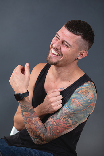 Studio portrait of a young tattooed man sitting in front of a black background wearing a tank top
