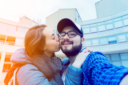 Young woman kissing partner on cheek while he smiles at camera very cheerfully
