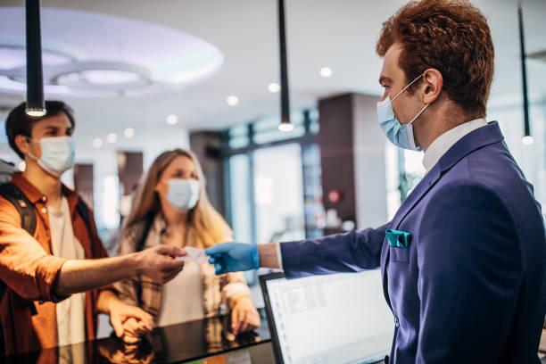 Couple travelers with medical masks on hotel reception talking to male receptionist Three people, young couple travelers with medical masks on hotel reception talking to male receptionist. concierge photos stock pictures, royalty-free photos & images