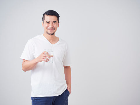 Handsome young man in a white T-shirt holding a glass of drinking water while standing in studio setting