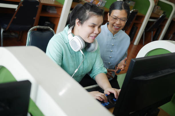 Asian young blind woman with headphone using computer with braille display assistive device discusing with senior colleague woman in workplace. Asian young blind woman with headphone using computer with braille display assistive device discusing with senior colleague woman in workplace. stock libraries stock pictures, royalty-free photos & images