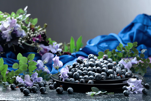 Fresh juicy blueberries on a black plate. Summer still life with blueberries, colored sweet peas and meadow grasses on a dark blue background.