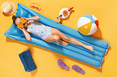 Little girl in a swimsuit and half glasses of orange resting on an inflatable blue mattress on a yellow background.