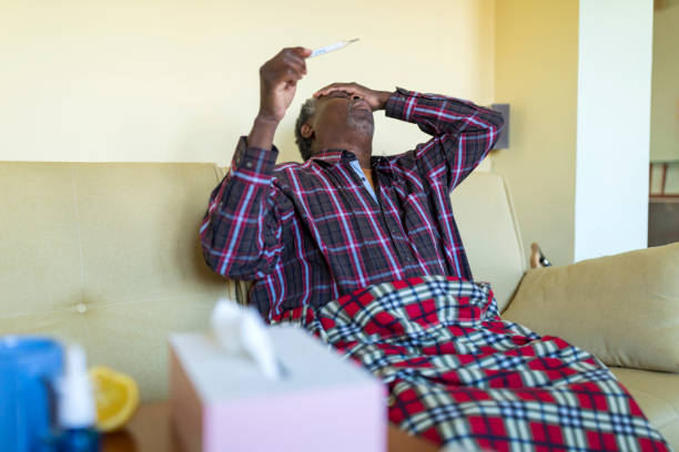 A senior Afro-American is sitting on the sofa at home with a bad health condition and looking at the thermometer and checking the temperature of his body. Ill African with seasonal infections, flu, allergy lying down in bed, covered with a blanket with high fever and flu.