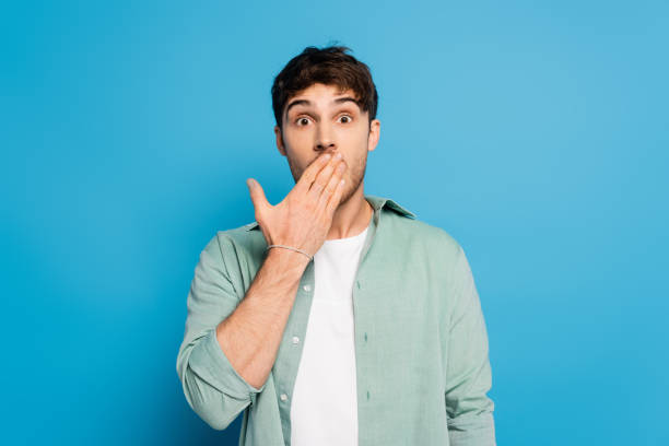 istock shocked young man covering mouth with hand on blue 1253998247