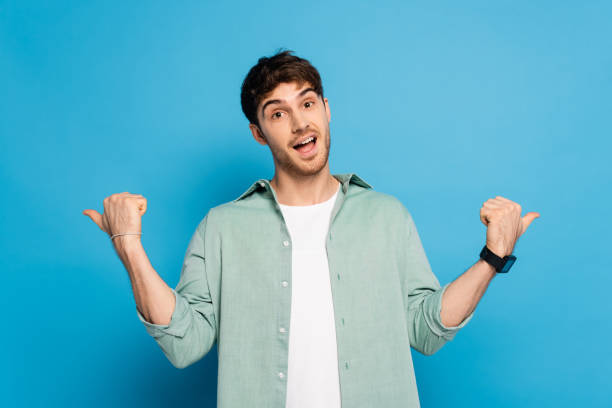 istock excited young man pointing with thumbs while looking at camera on blue 1253998249