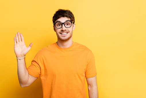 cheerful young man waving hand while looking at camera on yellow