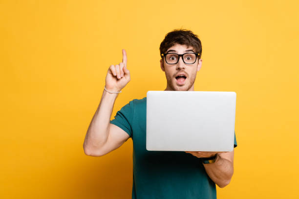 excited young man showing idea gesture while holding laptop on yellow excited young man showing idea gesture while holding laptop on yellow shocked computer stock pictures, royalty-free photos & images
