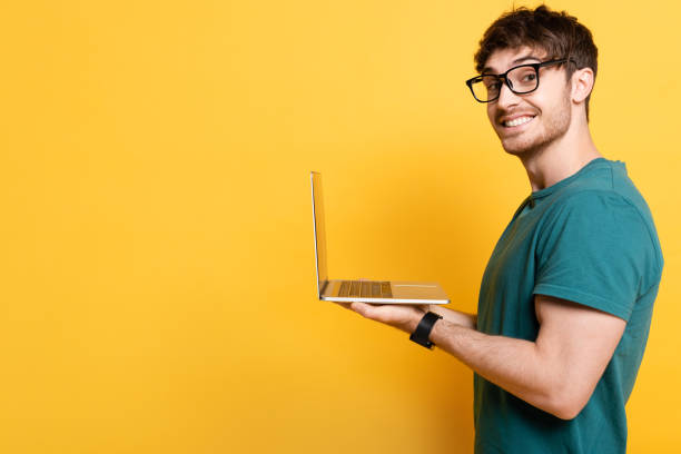 istock smiling young man looking at camera while holding laptop on yellow 1253998333