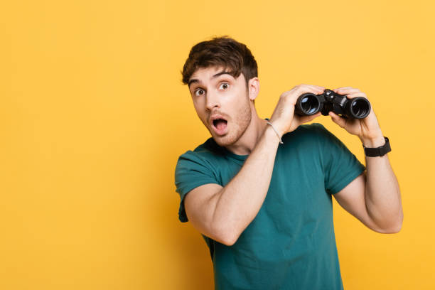 handsome surprised man holding binoculars and looking at camera on yellow