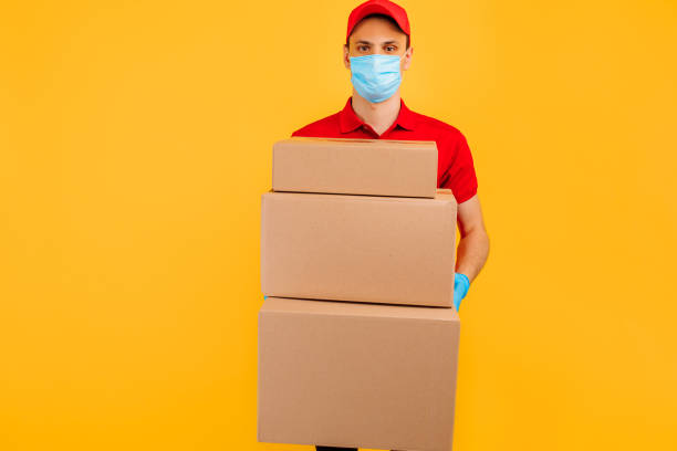 istock courier in a red uniform and a medical protective mask on his face holds cardboard boxes on an isolated yellow background. Online delivery, quarantine, coronavirus 1254006560