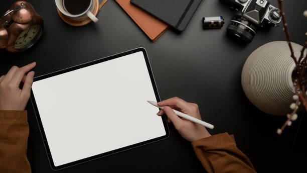 istock Female worker using blank screen tablet with stylus on dark luxury office desk with camera, supplies and decoration 1254009710