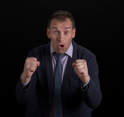 Portrait of excited businessman in suit with mouth open and clenching fists standing against black background.