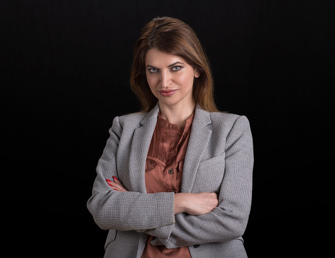 Portrait of businesswoman in grey blazer with arms crossed standing against black background.