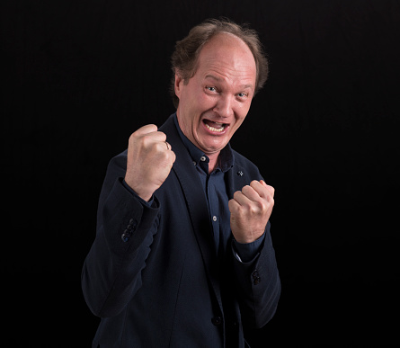 Portrait of excited mature man in blazer with mouth open and clenching fists standing against black background.