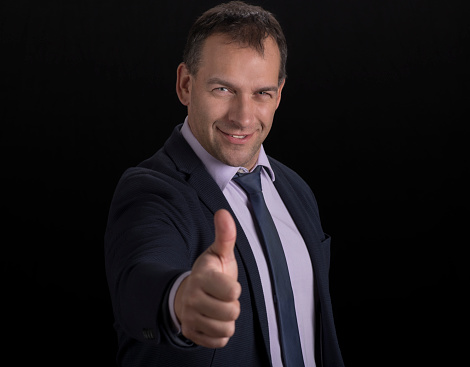 Portrait of smiling businessman in suit showing thumbs up sign standing against black background.