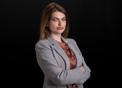 Portrait of businesswoman in grey blazer with arms crossed standing against black background.