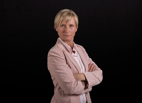 Portrait of serious businesswoman in pink blazer with arms crossed standing against black background.