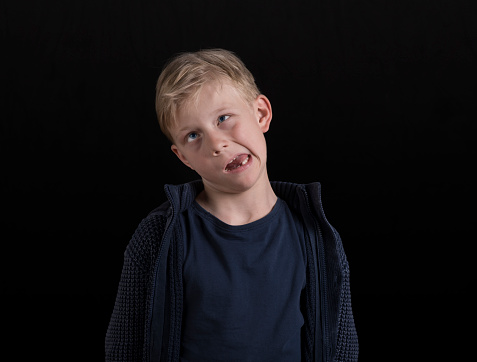 Portrait of boy in blue jacket pulling funny faces standing against black background.