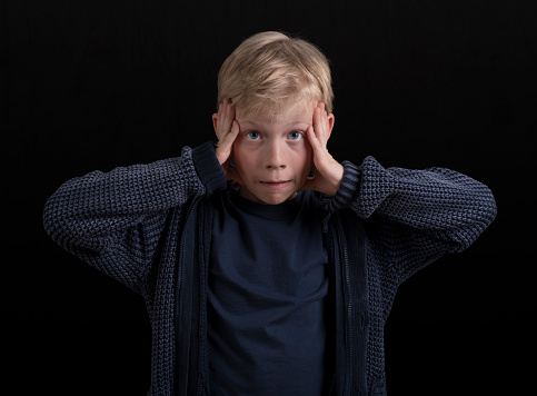 Portrait of frustrated boy in blue jacket with hands touching his head standing against black background.