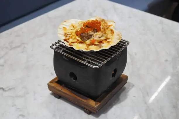 Photo of Grilled hotate or scallop spicy cheese lava with truffle and ebiko, served in  shell over grate and oven.