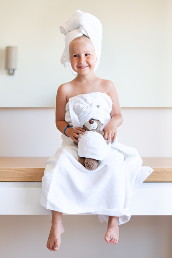 A little girl in a white bath towel and a turban  on her head sits on a table with her legs dangling, smiles, holds a teddy bear in her hands. Family look. Spa, after a shower, a beauty salon.