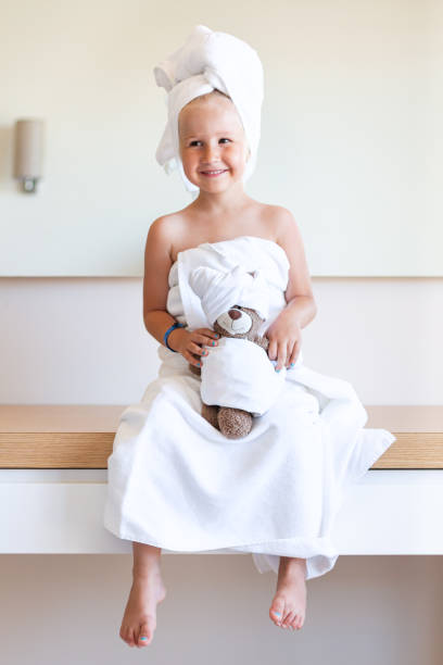 Little girl in a bath towel and a turban sits on a table with her legs dangling,smiles,holds a teddy bear.Family look.Spa,after a shower,beauty salon A little girl in a white bath towel and a turban on her head sits on a table with her legs dangling, smiles, holds a teddy bear in her hands. Family look. Spa, after a shower, a beauty salon. girls bath stock pictures, royalty-free photos & images