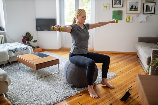 Woman sitting on a fitness ball in living room and working out with dumbbells. Woman is doing online exercise with digital tablet in front during self isolation at her living room.