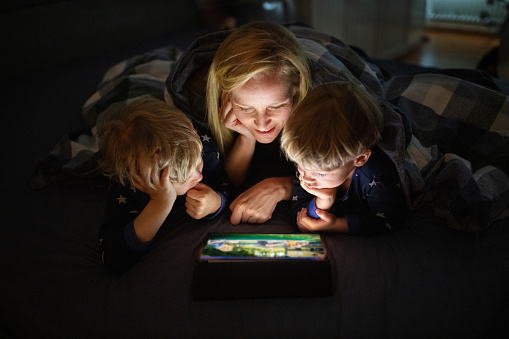 Mother and two sons lying on bed and enjoying watching movie on digital tablet. Mother and children using digital tablet before going to bed.