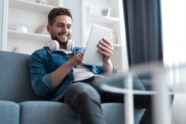 istock Attractive young man is using a tablet and smiling while lying on sofa at home. 1254047683