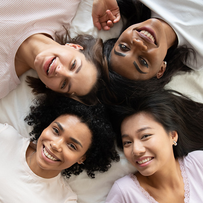 Top view diverse smiling young women best friends wearing pajamas relaxing lying in bed together, portrait from above beautiful girls looking at camera, having fun at hen-party, square photo