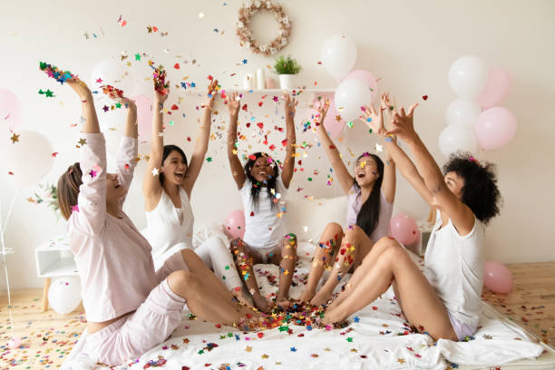 istock Happy diverse girls sitting on bed in circle, throwing confetti 1254049059