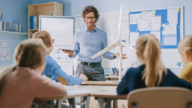 istock Elementary School Physics Teacher Uses Interactive Digital Tablet to Show to a Classroom of Smart Children how Renewable Energy and Wind Turbines Works. Science Class with Kids Listening Attentively 1254050833