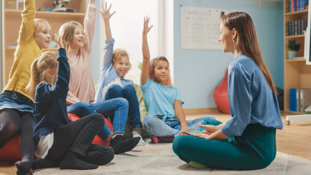 istock Elementary School Creativity Class: Children Sitting on the Bean Bags while Teacher Explains Lesson and Asks Question, Every Child Raises Hand with Answer. Learning in Modern Environment. 1254050979