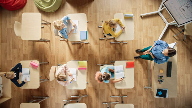 bright elementary school classroom: i bambini seduti al banco della scuola che lavorano, fanno un compito, insegnante entusiasta si alza un capo della classe spiegando una lezione. colpo di vista dall'alto. - top of the class foto e immagini stock