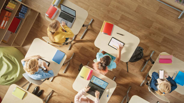 scatto in alto nella classe di informatica della scuola elementare: bambini seduti alla scrivania della scuola utilizzando personal computer e tablet digitali per compiti. - educazione foto e immagini stock