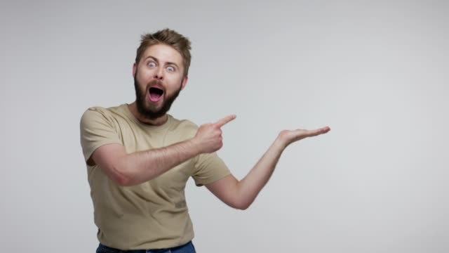 Surprised bearded guy pointing aside, holding empty space on palm and looking with open mouth amazed