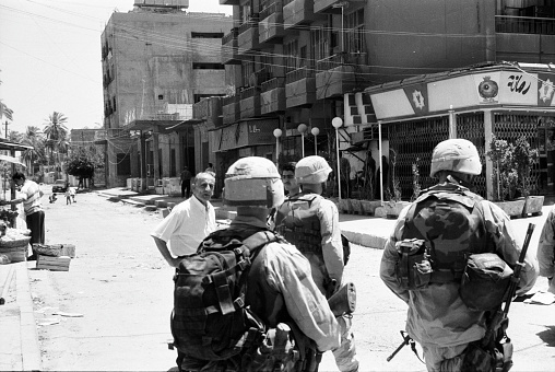 A troop of U.S. soldiers is surveilling the streets of Baghdad. There was a lot of chaos there in 2003 due to criminals and pendants of Saddam Hussein. The man in the background watches the soldiers suspiciously. Photographed by Michael Multhoff in Baghdad / Iraq. May 12, 2003