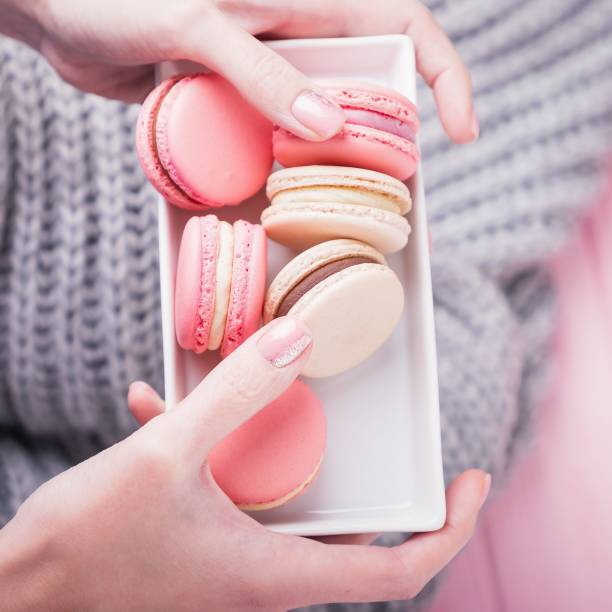 istock Female hands holding rectangular white plate with delicious pink and beige macaroons on pink and gray background. Top view 1254053765