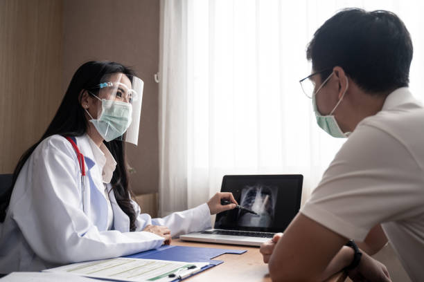 le docteur féminin expliquent le résultat de coffre de x-ray dans l’ordinateur portatif à patient d’homme de maladie dans la salle d’examen à l’hôpital. les femmes medic portent un bouclier et un masque pendant la pandémie de covide pour pré - résultat dexamen photos et images de collection