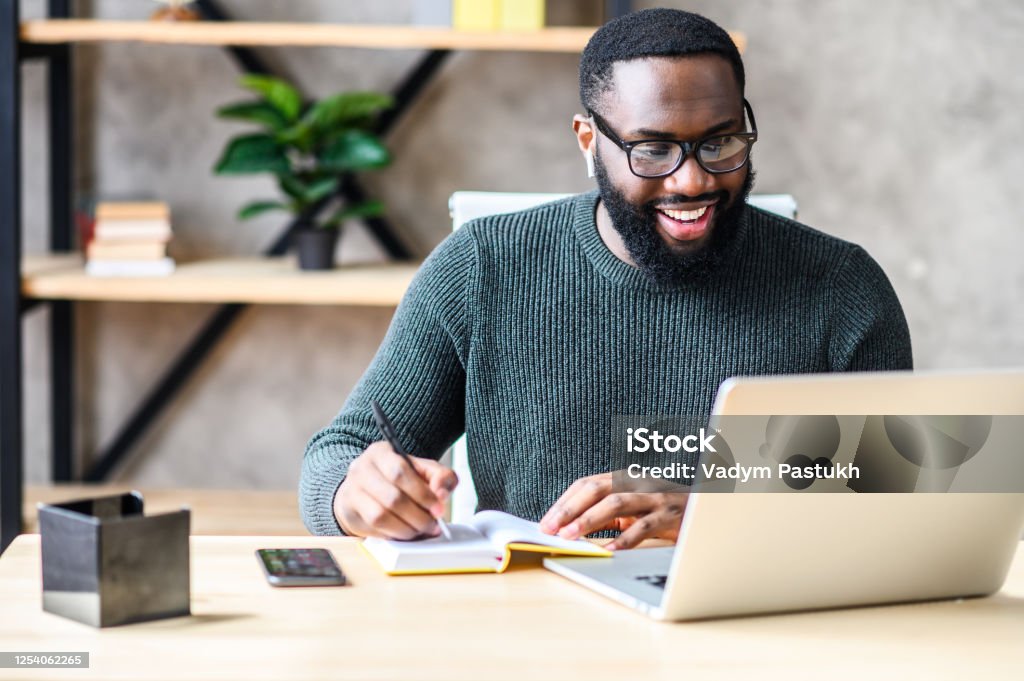 An African-American guy using laptop in the office Cheerful African-American male student or worker in glasses is watching online lectures or webinars and writing notes in a notebook Men Stock Photo