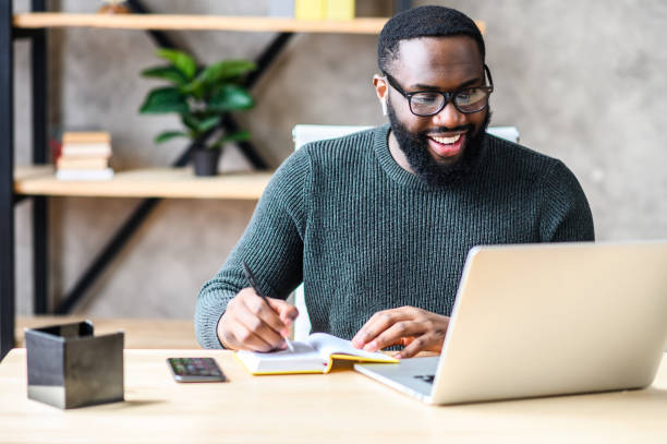 An African-American guy using laptop in the office Cheerful African-American male student or worker in glasses is watching online lectures or webinars and writing notes in a notebook conference call stock pictures, royalty-free photos & images