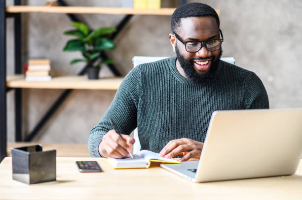 An African-American guy using laptop in the office Cheerful African-American male student or worker in glasses is watching online lectures or webinars and writing notes in a notebook student desk stock pictures, royalty-free photos & images