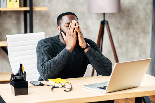 Exhausted African-American office employee sits at the desk with a laptop and covers face with his hands, tired guy closed eye