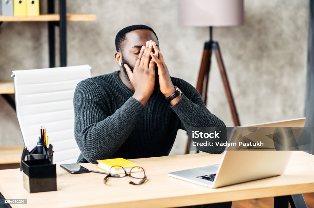 Tired African-American sits at the desk Exhausted African-American office employee sits at the desk with a laptop and covers face with his hands, tired guy closed eye Frustration Stock Photo